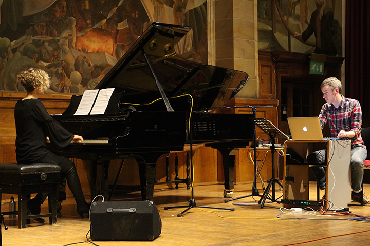 A photo of concert pianist Xenia Pestova Bennett on stage playing the piano with electro-acoustic artist Ed Bennett playing the interludes on a laptop.