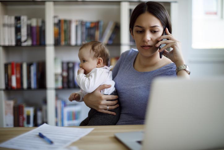 A colour photo of a woman wearing a blue top at a desk on the phone holding a baby in a white babygro sucking its thumb.