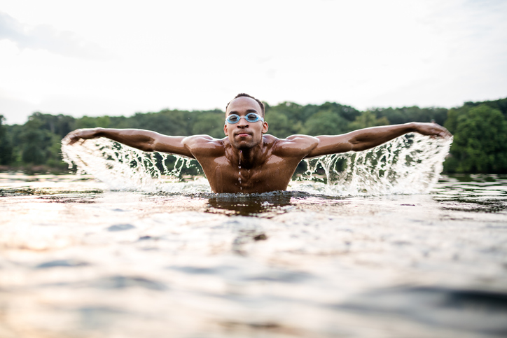 A photo of a black man wearing swimming goggles doing butterfly stroke in a lake with trees in the background. 