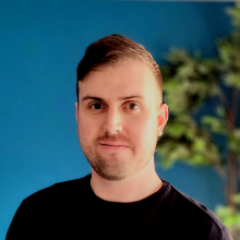 Headshot of Adam standing against a blue wall. Adam has light brown hair and is wearing dark clothing.