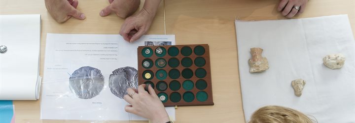 Downward shot of a table scattered with coins, bones and pieces of paper with four people reaching across from just outside the photo edges