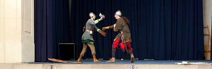 Photograph of two figures in Viking clothes fighting on stage in front of an audience of schoolchildren.