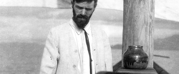 Black and white photographic portrait of DH Lawrence in a white suit jacket stood next to a wooden mast.