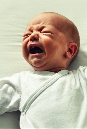 A baby wearing a white longsleeved outfit crying, lying on a white surface