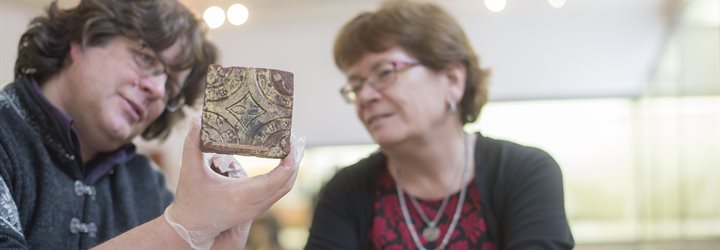 Dr Roderick Dale and Professor Judith Jesch sat at a table in the museum looking at a floor tile from a medieval abbey in Shropshire, Derby Museum and Art Gallery