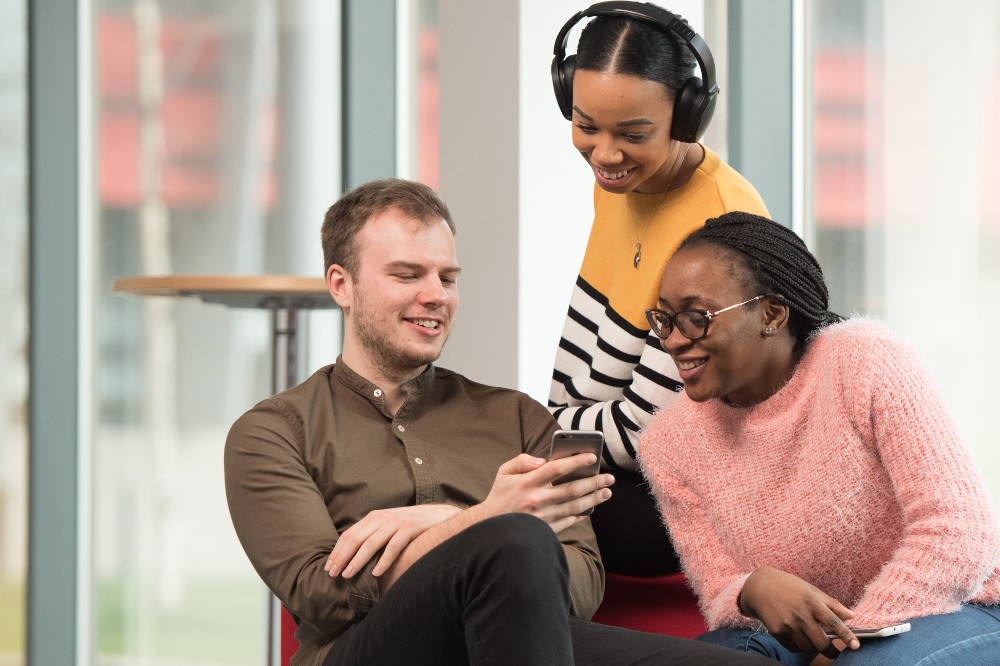 Three postgraduate students looking at a phone with one wearing headphones