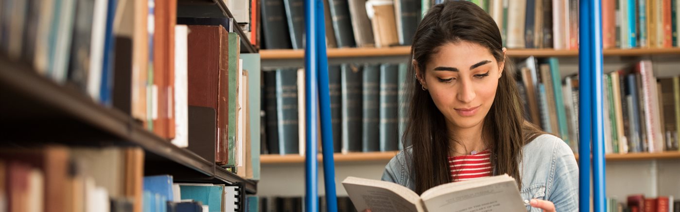 Student reading book while standing on library ladder surrounded by bookshelves