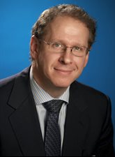 Photo of David Falzani. David is smiling and wearing glasses. he is wearing a dark jacket and dark tie with a pale striped shirt.