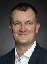 Head and shoulders photo of Paul Riseborough. Paul is wearing a dark jacket with a white shirt and is smiling.