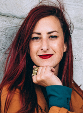 Head and shoulders photo of Vanja Ljevar. Vanja has long burgundy hair and is wearing an orange shirt with green cuffs. Her hand is resting under her chin and she is smiling.