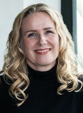 Head and shoulders photo of Nichola Elenor. Nichola is smiling and has long wavy blond hair. Nichola is wearing a high neck black jumper.