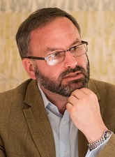 Head and shoulder photo of Richard Profit. Richard is wearing glasses, has a beard and has his hand resting against his chin. He is wearing a light brown jacket and a pale shirt.
