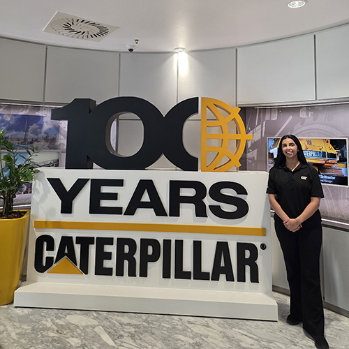 Student Ankia Dhunna, stands beside a large Caterpillar Inc. centennial display. The installation features a bold "100" logo with the final zero designed as a yellow globe outline. Beneath it, large block lettering reads "YEARS" and "CATERPILLAR".