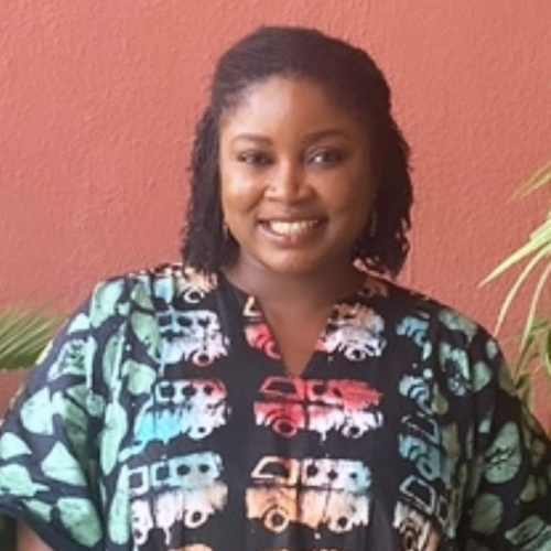 Janet Torhee smiling, with short black curly hair wearing a bright batik printed top, standing in front of a terracotta wall