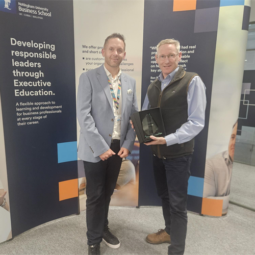Photo of Mark Boardman receiving an award for excellent engagement from Executive Education staff member Nick Proverbs at the end of the Help to Grow: Management course. Mark holds the award and they both smile at the camera