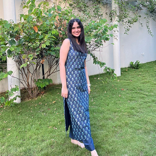 Serah Dominic standing in front of some tall bushes on grass, wearing a dark blue printed dress, has long dark hair and smiling.