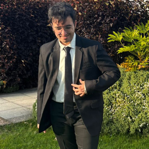 Shubham Thakkar smiles, with short black hair and glasses, wearing a black suit and tie over a white shirt, standing on grass in front of bushes.