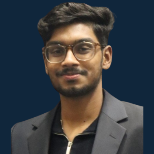 Thejeshwar Karthikeyan with short dark hair and beard, wearing glasses and smiling. Wearing a dark grey blazer and black zip top