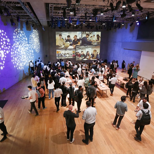 A photo of an event indoors with lots of people in a room with large projected images on the wall. The photo is taken from a high up angle, looking down across the room.