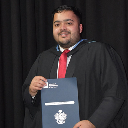 Umang Agrawal wearing a black graduation gown with a red tie, holding a certificate standing in front of a black curtain