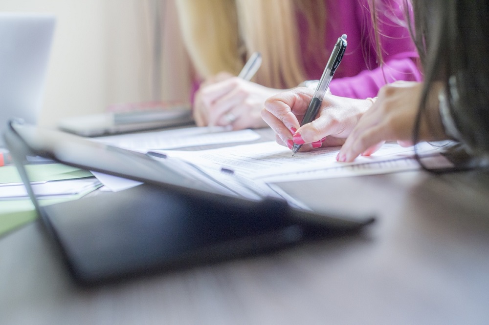 Close-up of two people's hands holding a pen and writing whilst sat at a table with a laptop on it in the foreground.