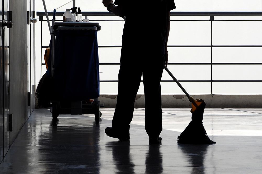A black and white photo showing a domestic worker mopping a floor. The worker is visible from the waist down only and is in silhouette. They are stood next to a cleaning trolley which is also in silhouette.