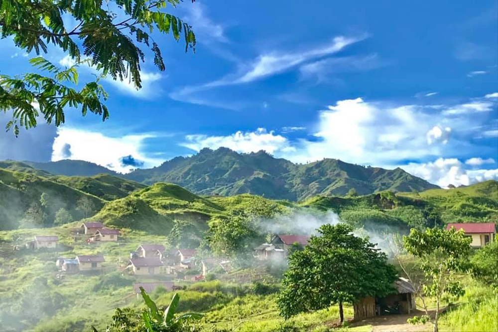 A landscape view of a remote village on the island of Mindanao, Philippines. The scene captures sunny weather with patches of lingering clouds, suggesting a recent storm has passed. The village appears nestled within lush greenery.