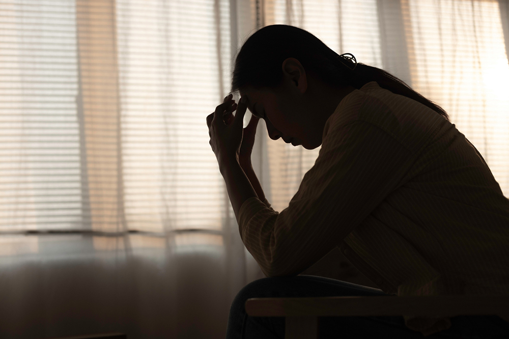 Silhouette of a person sitting indoors with head bowed and hands resting on the forehead, positioned in front of softly lit curtains.