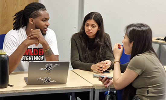 Three students sitting at a table with a laptop in front of them and talking