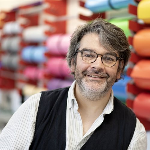 Tom Hignett, a Graphic Design Technician/ Demonstrator from the University of Chester, stands in a design studio. He is dressed in smart-casual clothes and smiles at the camera. Behind him, rolls of coloured vinyl are mounted on the wall.