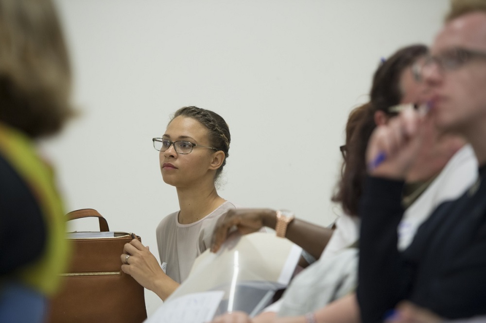 Mature students in a seminar room. In focus is a female student who is wearing glasses with her hair tied back.