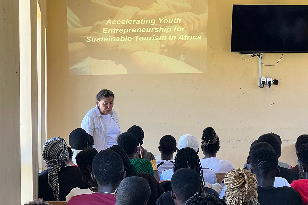 Photo of Professor Marina Novelli delivering a lecture in a classroom setting with 'Accelerating youth entrepreneurship in tourism for sustainable development' words projected on the wall. In the foreground are students sat down with their backs to the camera.