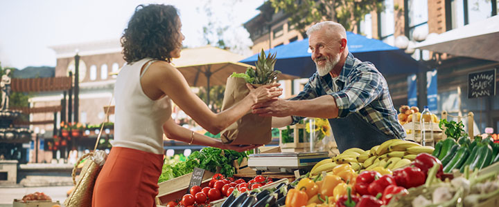 Street vendor running a small market stall selling fresh locally produced fruit and vegetables. He is serving a customer filling a recycled paper bag to put the produce  in.