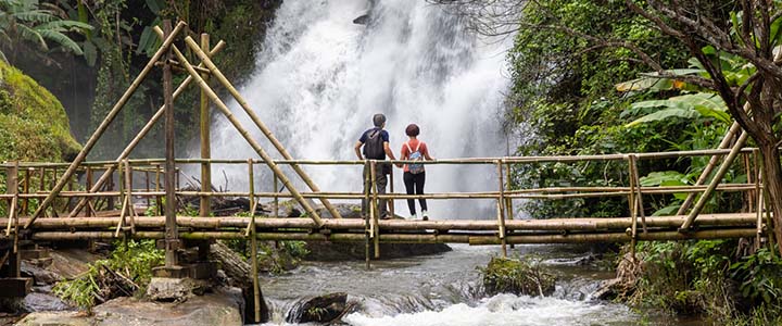 A couple stood on a wooden bridge in a tropical forest looking at a waterfall that flowers under the bridge.