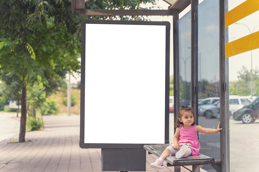 A blank digital billboard at a bus stop. A young child is sitting at the bus stop wearing a pink dress.