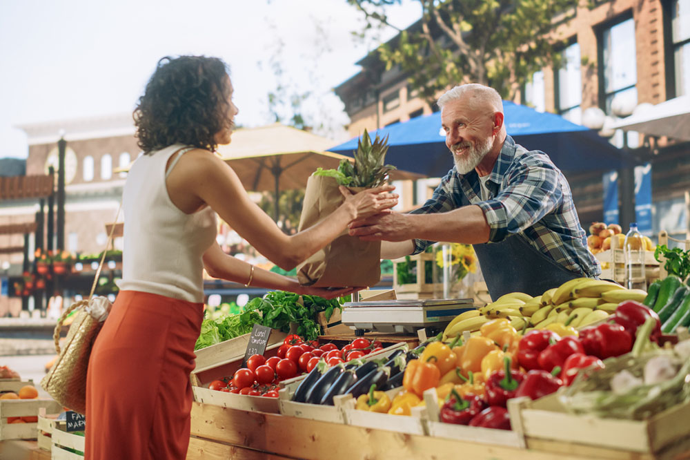 A street vendor running a small farm market business, selling sustainable fruits and vegetables. He is serving a customer and filling a recycled paper bag with fresh produce.