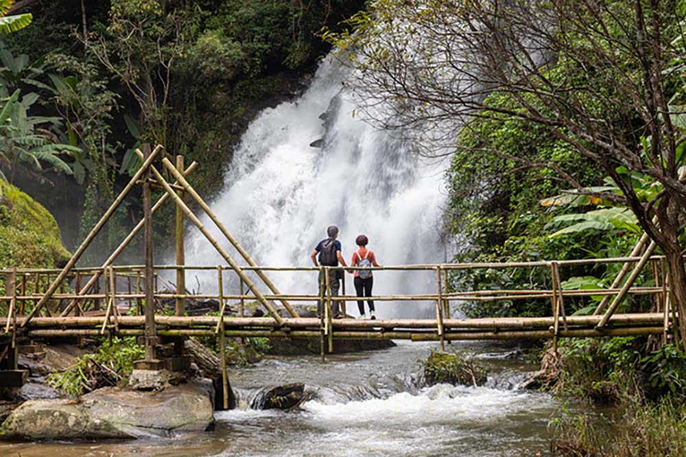 A couple standing on a wooden bridge in a tropical forest looking at a waterfall that flows under the bridge.