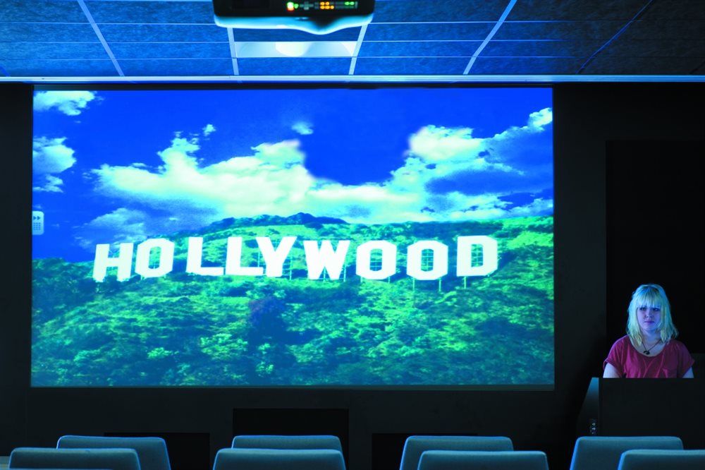 Person standing behind lectern at front of cinema with Hollywood sign displayed on screen