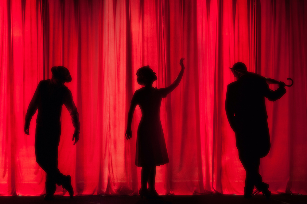 The silhouette of three performers can be seen through a red curtain
