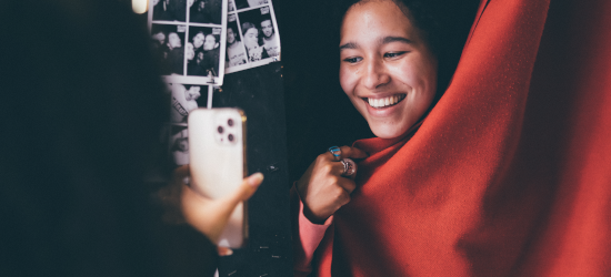 Student taking photo on phone of friend peering out from behind curtain, smiling