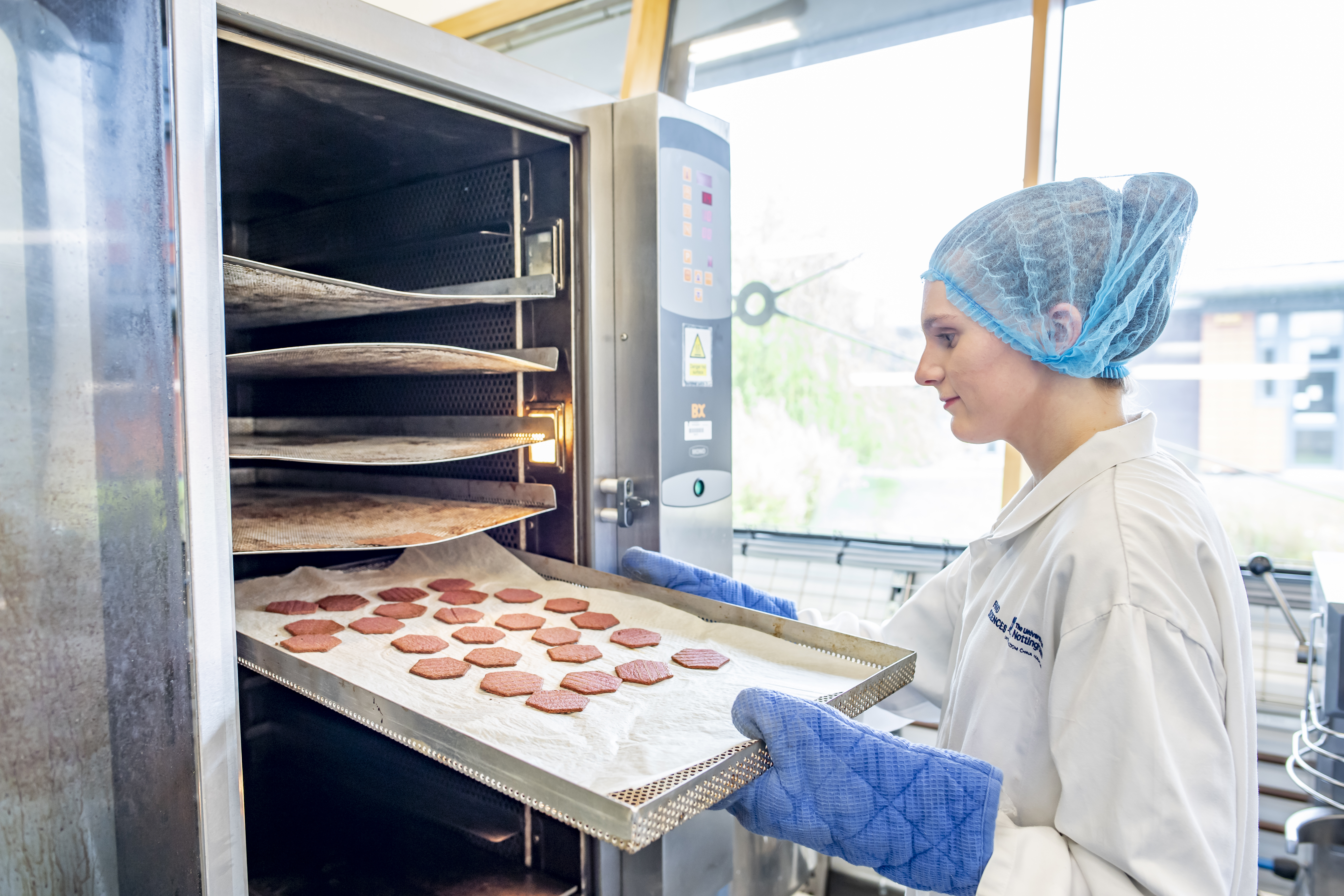 Undergraduate student Katherine Tolson taking baked products from an oven wearing lab coat, hair net and oven gloves