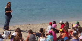 A large group of children seated on the beach listening to a woman who stands with her back to the ocean with several islands in the distance, beneath clear blue skies (photo).