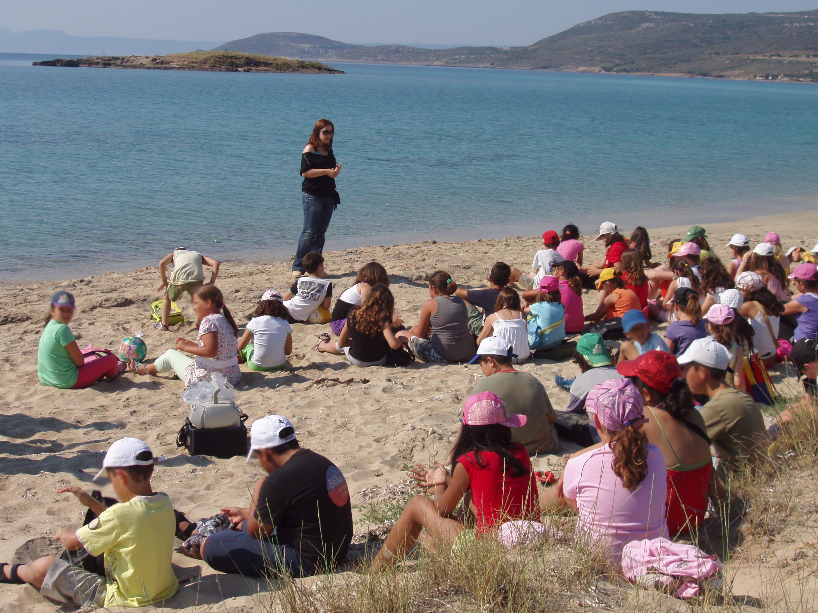 Image shows a large group of children seated on the beach listening to a woman who stands with her back to the ocean with several islands in the distance, beneath clear blue skies