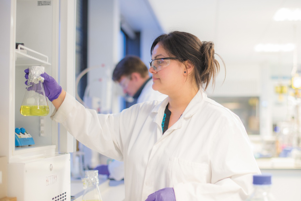 Female researcher in lab coat and gloves, inspecting up a chemical flask