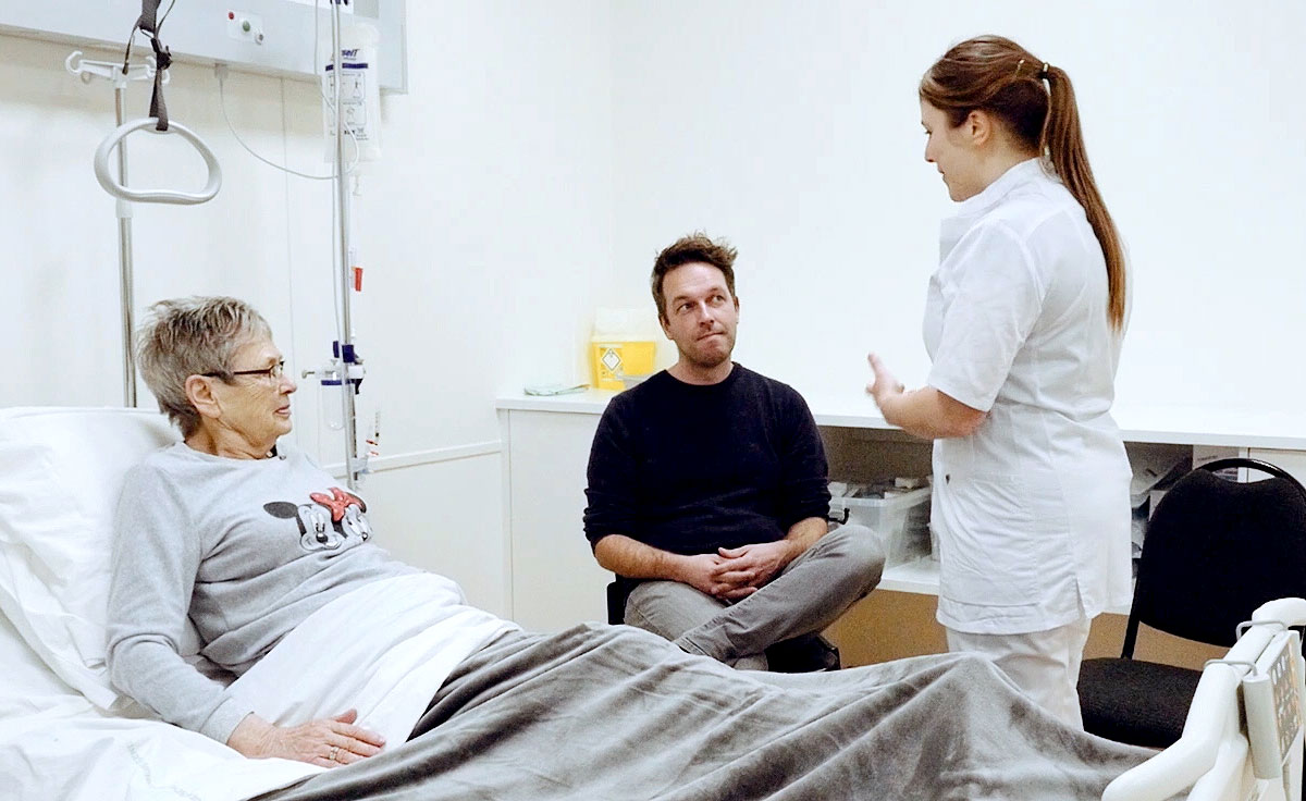 Video still 1 - Scene showing patient in bed in a hospital setting. The Nurse and Carer are at the bedside.