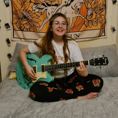 Amber Frost sitting cross-legged on her bed, smiling and holding a blue guitar
