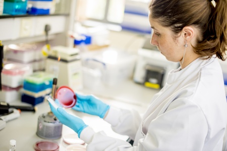 Female student wearing a lab coat and gloves looking into a red petri dish