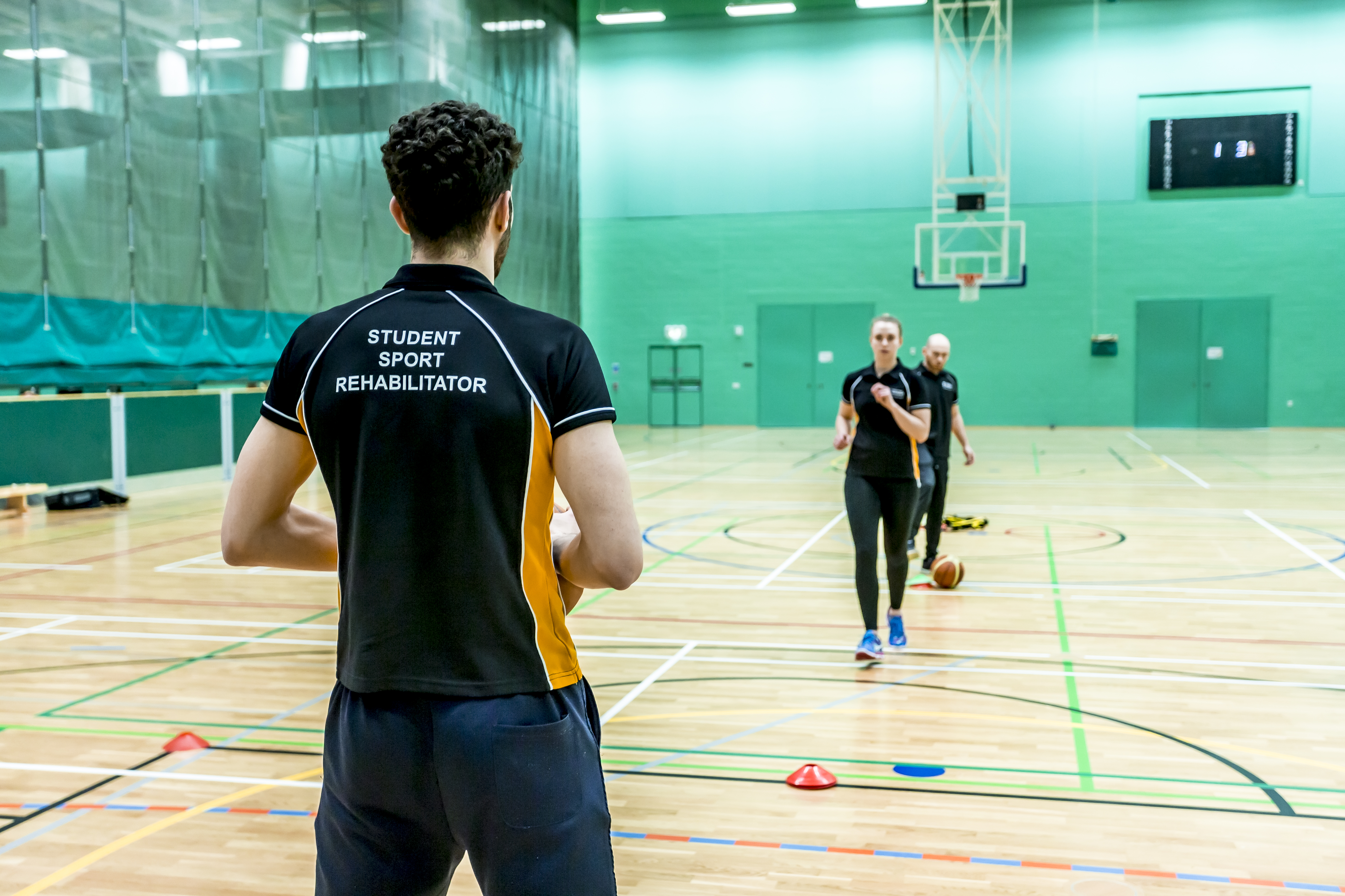 Student wearing a sport rehabilitation top in a gym sports hall coaching people who are running in gym clothes