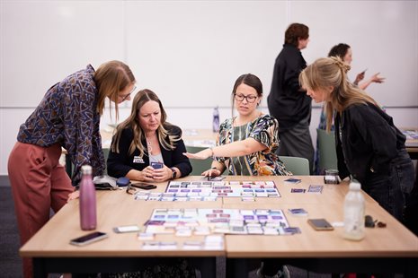 Four white women having a discussion around a table with a board game spread across the table