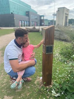 A man holds a girl as they press the button on the listening post in front of Lakeside Arts.